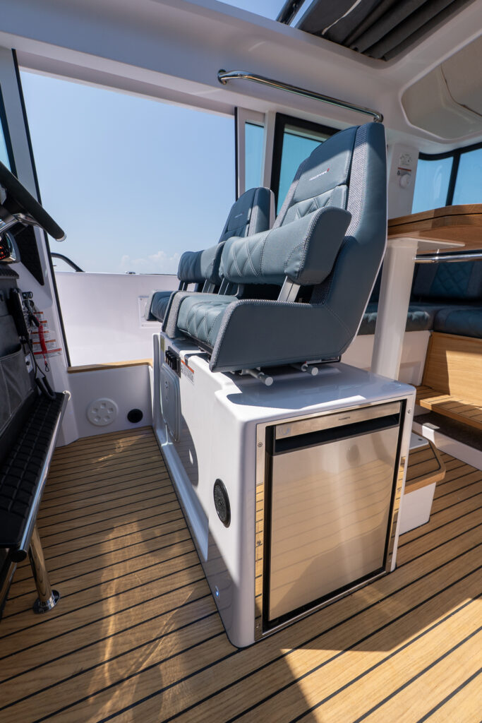 Twin bolster helm seats on an Axopar 37 mounted above an integrated stainless steel refrigerator, with teak flooring and cabin lounge visible in the background.