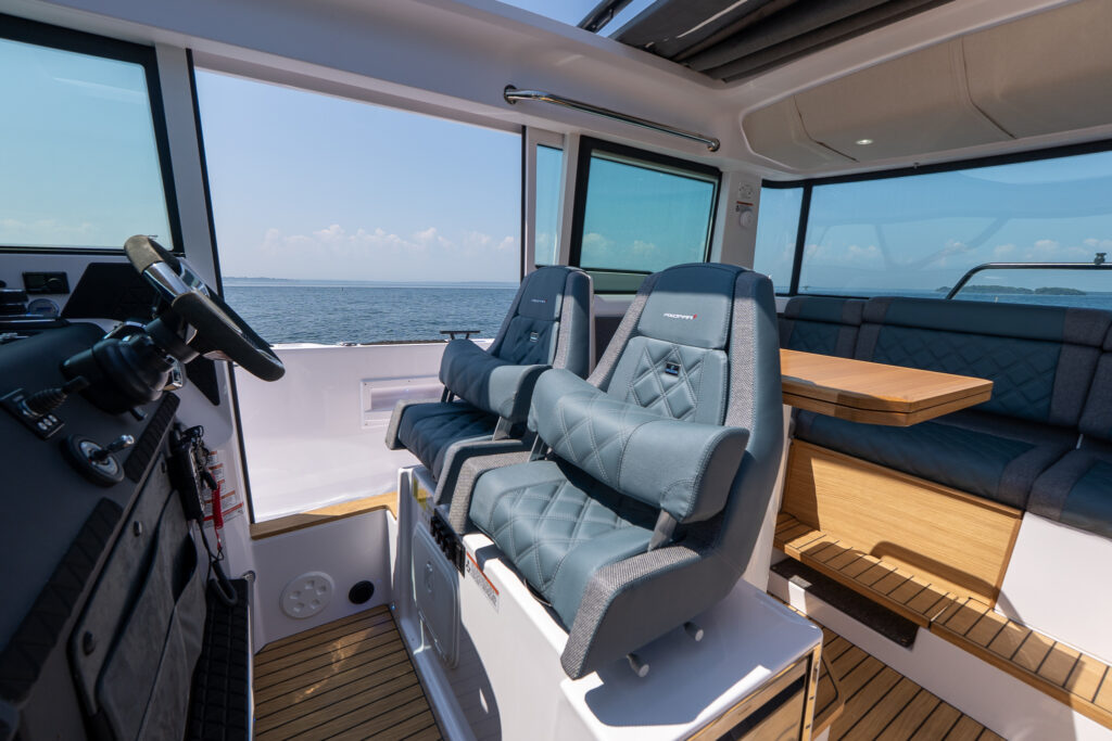Twin bolster helm seats on an Axopar 37 facing the steering console, with an L-shaped aft lounge and teak table visible through large cabin windows overlooking open water.