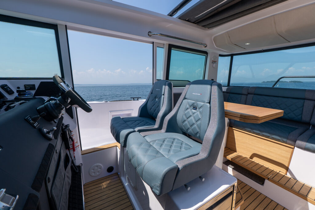 Twin diamond-stitched helm seats on an Axopar 37 facing the steering console, with an aft lounge and teak table visible through large cabin windows overlooking open water.
