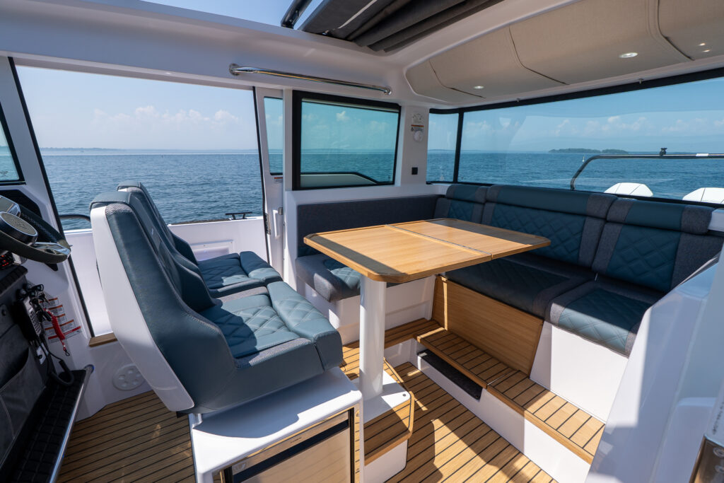 Interior seating area of an Axopar 37 featuring twin helm seats and a large L-shaped lounge with a teak dining table, surrounded by wide cabin windows overlooking the water.