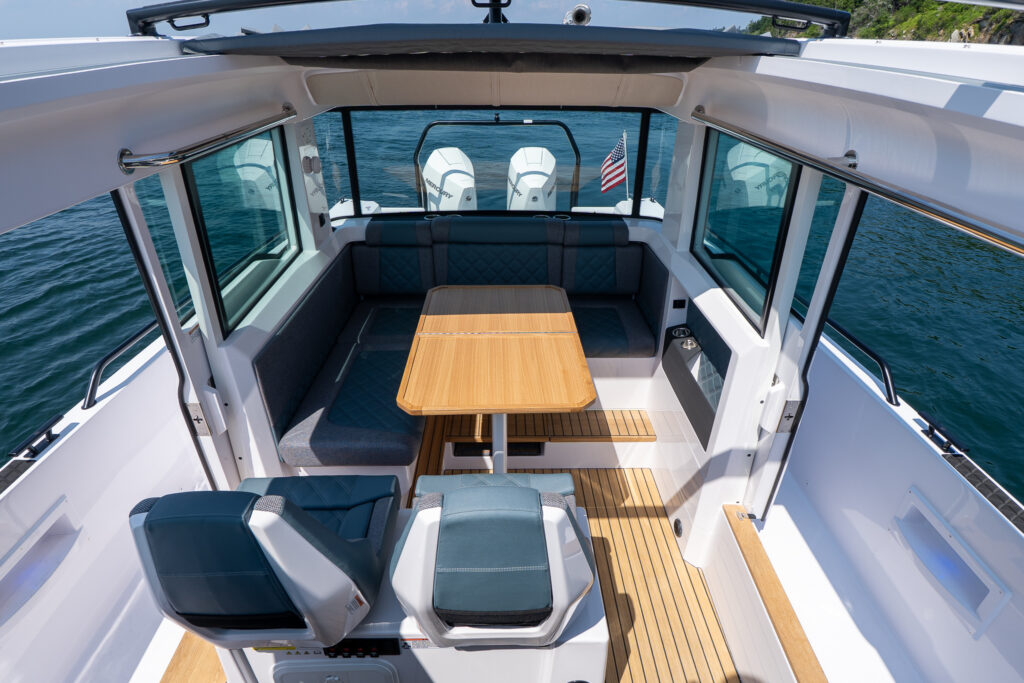 Top-down view of the Axopar 37’s interior showing twin helm seats, a teak dining table, and a U-shaped aft lounge with diamond-stitched cushions, framed by large windows and an open roof.
