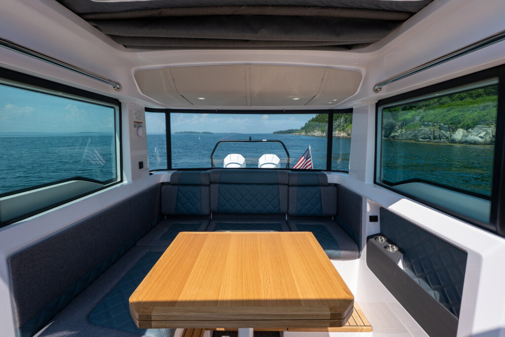 Aft cabin seating area on an Axopar 37 featuring a U-shaped lounge with diamond-stitched cushions, teak dining table, and wraparound windows overlooking the water and coastline.