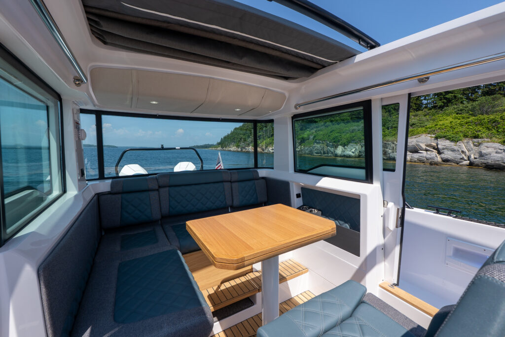 Aft cabin lounge of an Axopar 37 featuring a U-shaped seating area with diamond-stitched cushions, a teak dining table, large side windows, and views of the surrounding coastline.