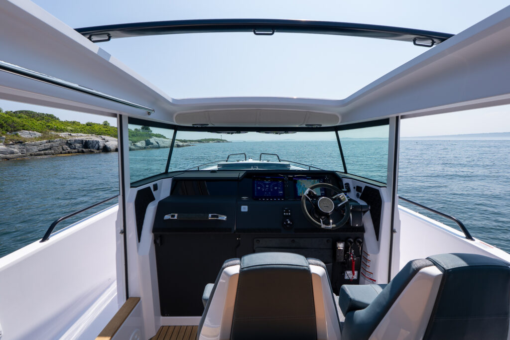 Helm view from inside an Axopar 37 showing twin multifunction displays, a premium steering wheel, open roof panels, and panoramic views of the surrounding coastline and open water.