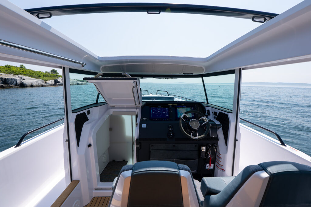 View from the helm seating of an Axopar 37 showing the steering console with dual displays, the open cabin entry hatch, and expansive ocean views through the wraparound windows and open roof.