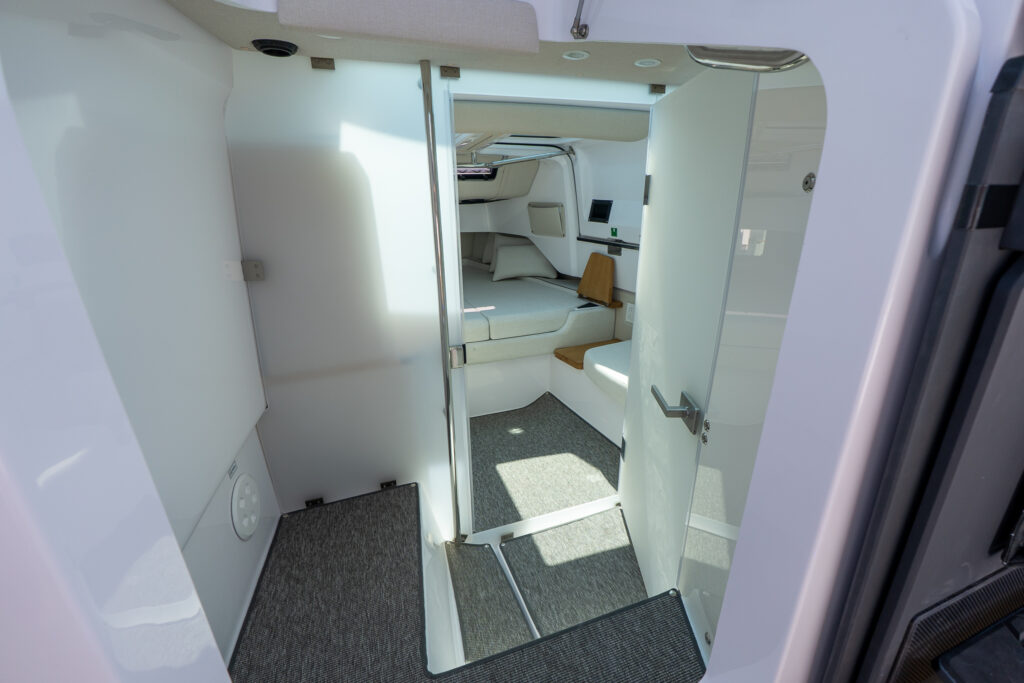 View through frosted glass doors into the forward cabin of an Axopar 37, showing the cushioned berth, side shelving, and soft interior panels with carpeted flooring.