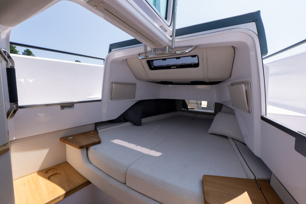 Bright forward berth on an Axopar 37 with cushioned sleeping area, overhead hatch, side storage pockets, and wooden shelving, viewed from the open cabin entry.