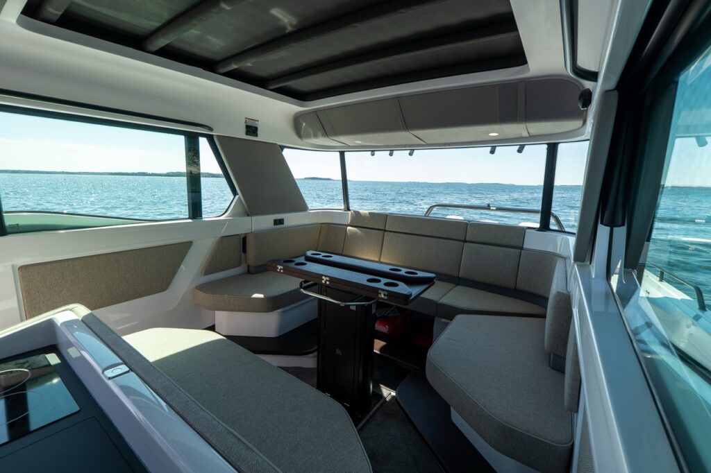Interior view of the Axopar 45 cabin showing wraparound aft lounge seating in Sandstone upholstery with a central table and wide panoramic windows overlooking the water.