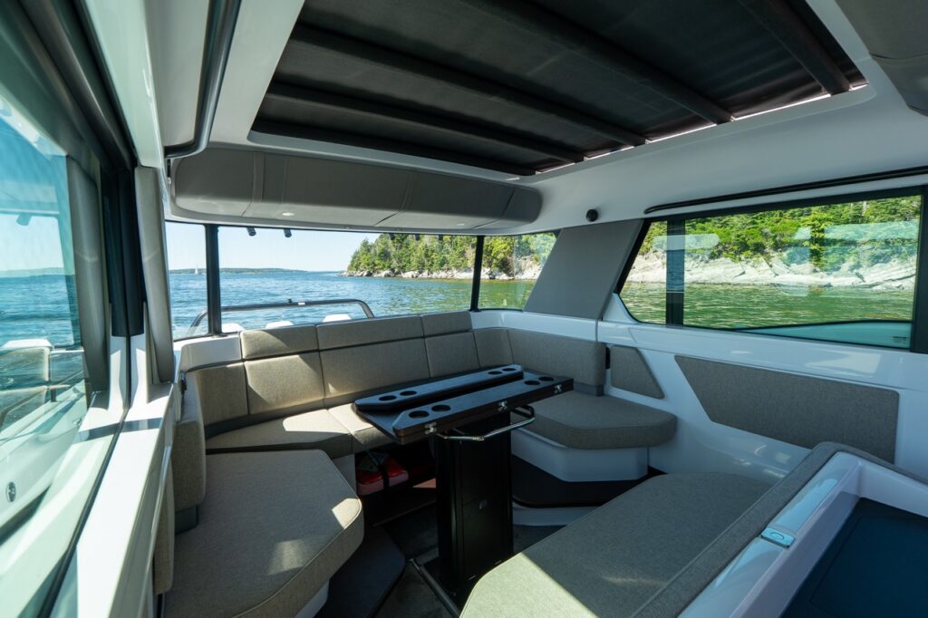 Cabin interior with wraparound lounge seating in Sandstone upholstery, a pedestal table with cupholders, and large windows framing a rocky, tree-lined shoreline.