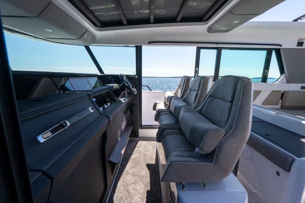 Three upholstered pilot seats face forward beside the helm console, where twin Multi Function Display (MFD) screens sit under the windshield with the side door open to the sea.
