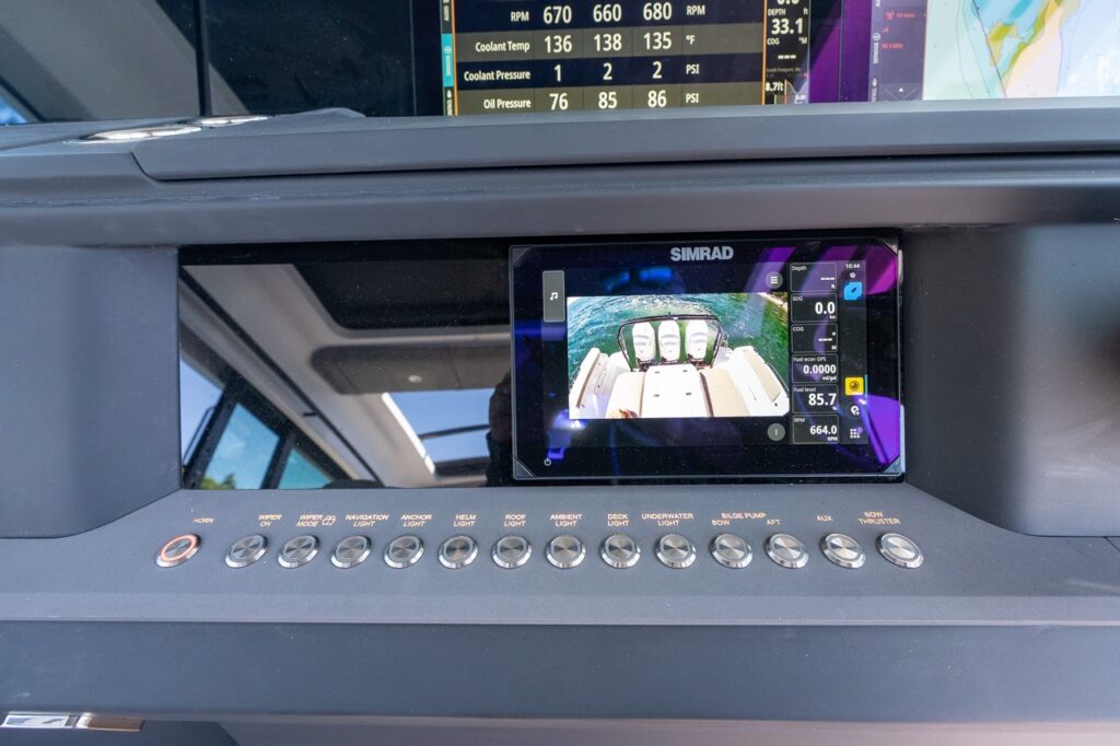 Close-up of a Simrad Multi Function Display (MFD) showing a rear camera view of triple outboards, mounted above a row of stainless push-button switches labeled for wipers, navigation lights, anchor light, helm light, roof light, deck light, underwater lights, bilge pump, and bow thruster.