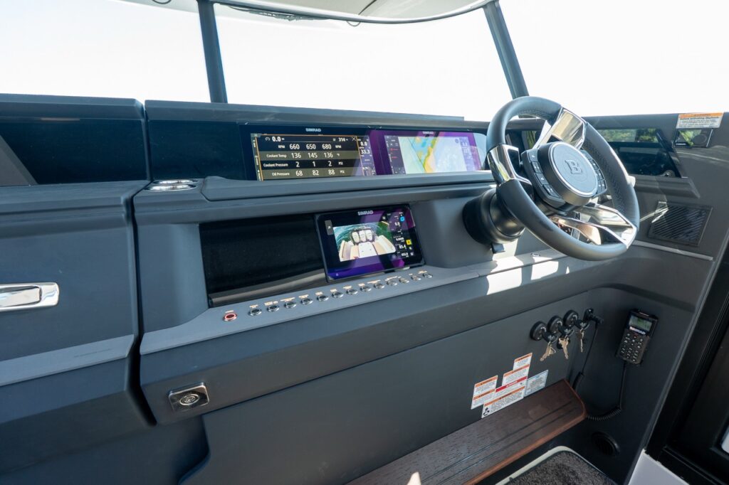 Helm of an Axopar 45 showing dual wide Simrad Multi Function Displays, a lower Simrad camera/navigation screen, push-button switch row, steering wheel, throttle controls, and mounted VHF handset.