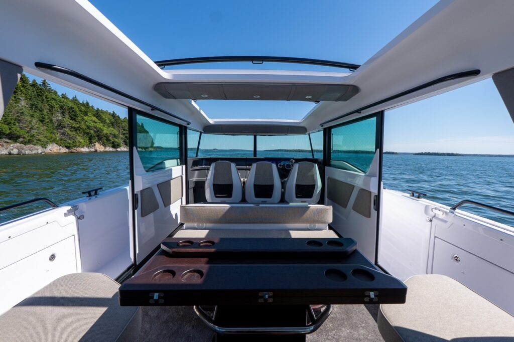 View forward from the aft seating area into the pilothouse with the rear pilothouse doors open, both sunroofs open overhead, and the cabin table in the foreground with three helm seats ahead and water on both sides.