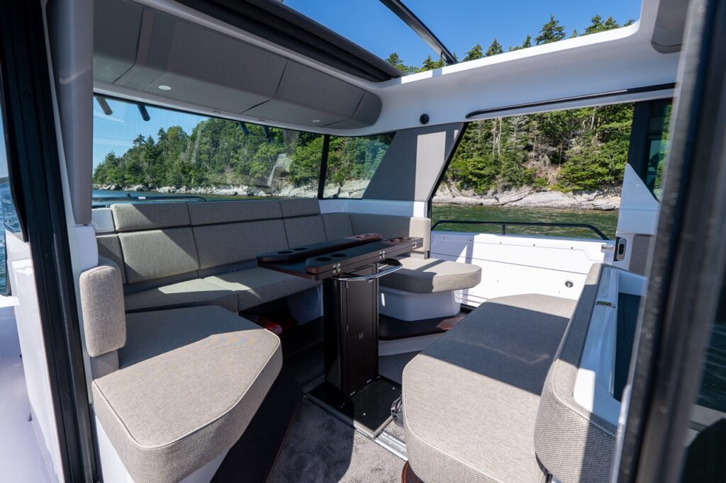 Inside the pilothouse lounge, Mediterrana upholstery wraps around a U-shaped settee beside a pedestal table, with the starboard side door open to the shoreline and sunlight streaming in through the open sunroof.