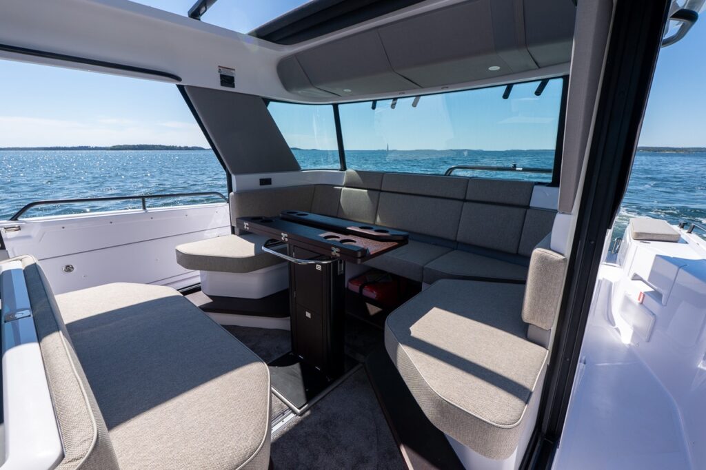 Inside the pilothouse, Mediterrana-upholstered L-shaped seating wraps around a pedestal table with cupholders, with side doors open to the sea view.