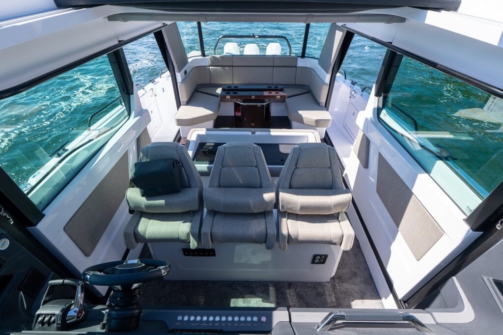 Wide view from the helm looking aft through the pilothouse, showing three forward-facing seats, the galley module behind them, and Mediterrana-upholstered lounge seating with table at the rear.