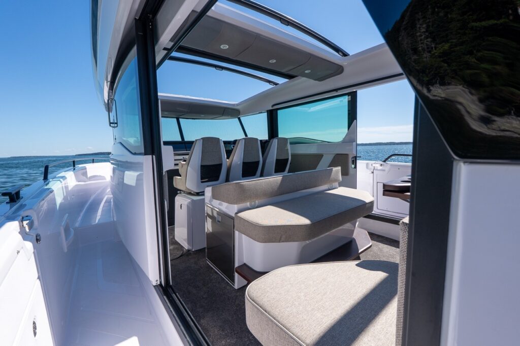 View into the pilothouse through an open side door, showing three helm seats, a slim aft-facing bench in Mediterrana upholstery, and both sunroofs open above with ocean visible outside.