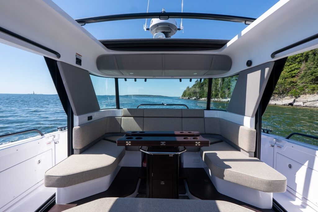 Interior view of the Axopar aft cabin lounge featuring Mediterrana upholstery, a central folding table with cupholders, wraparound seating, and both sunroofs retracted to reveal an open sky and ocean backdrop.