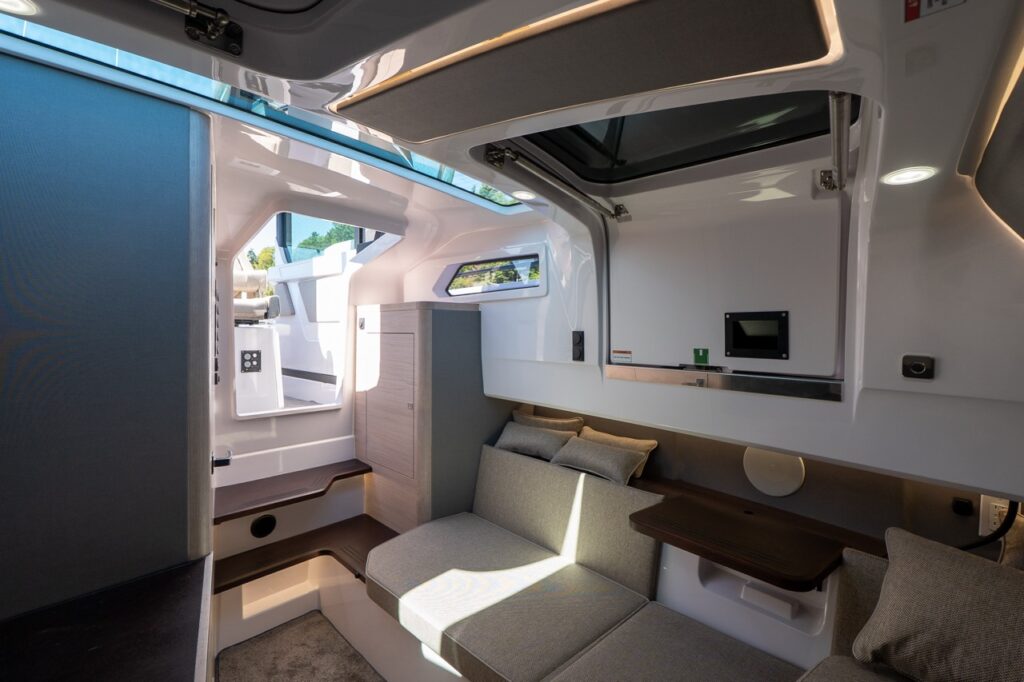 View inside the Axopar 45 forward cabin showing a cushioned lounge/berth in Mediterrana upholstery, wood-finish cabinetry, a side shelf table, integrated lighting, and overhead hatch bringing in daylight.