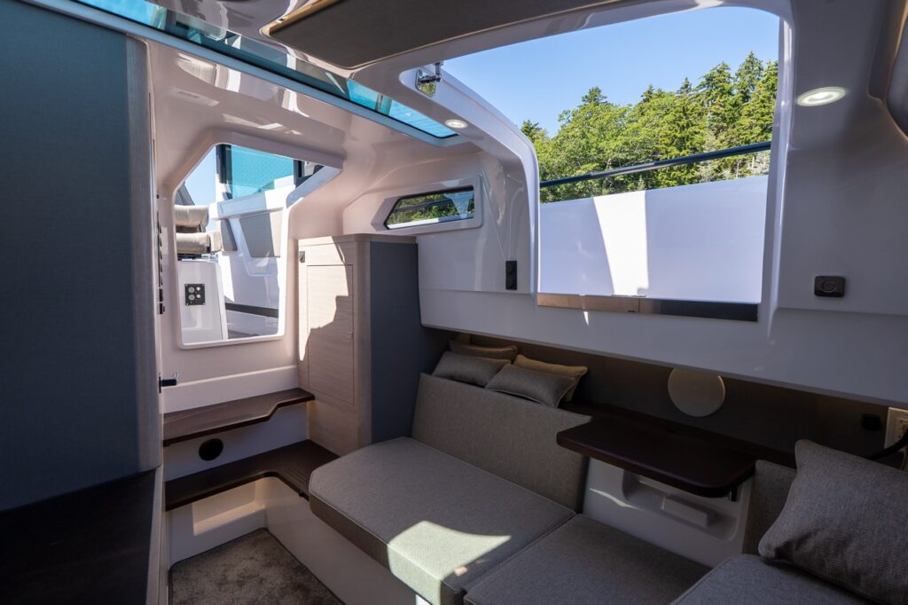 Inside the Axopar 45 forward cabin, sunlight pours in through an open overhead hatch above a Mediterrana-upholstered lounge/berth, with wood-finish storage, molded steps, and a small side table.