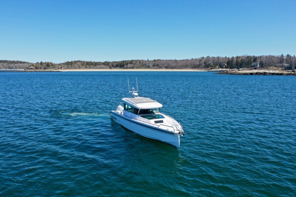 Aerial view of an Axopar 37 XC Cross Cabin cruising on calm blue water with a low, tree-lined shoreline in the distance under a clear sky.