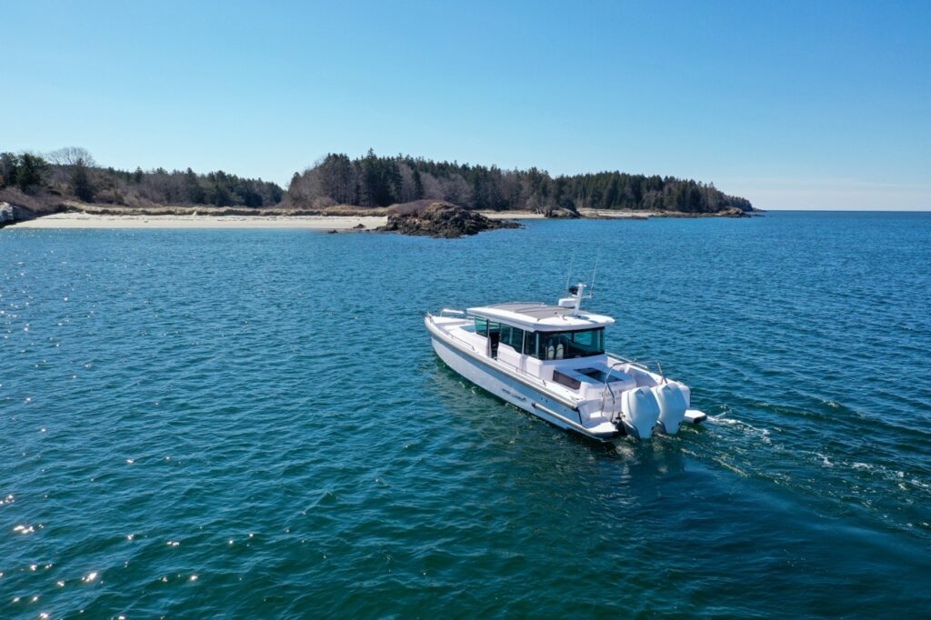 Drone view of an Axopar 37 XC Cross Cabin with twin outboards cruising on blue water near a sandy, tree-lined shoreline on a clear day.
