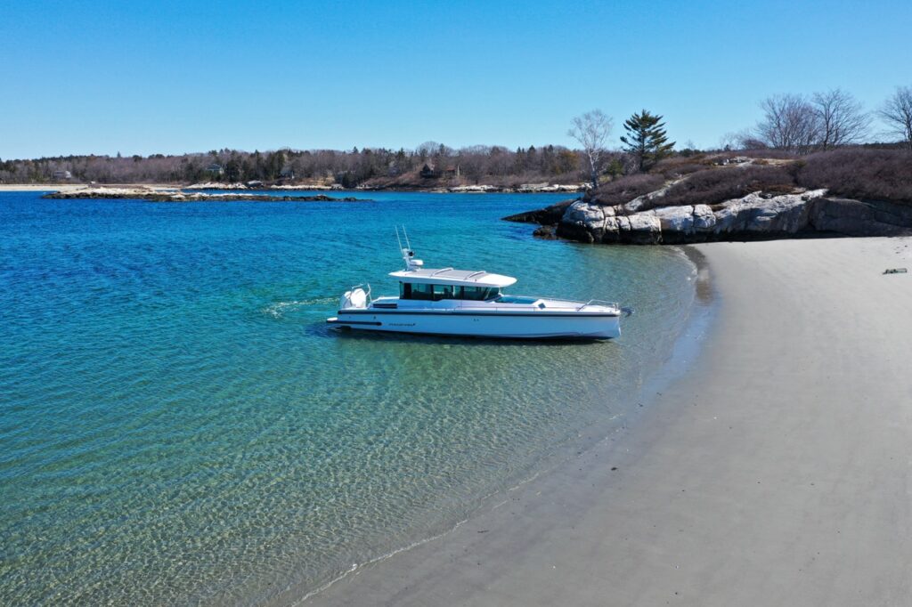 Drone side view of an Axopar 37 XC Cross Cabin positioned just off a sandy beach in clear shallow water, demonstrating safe beaching technique.