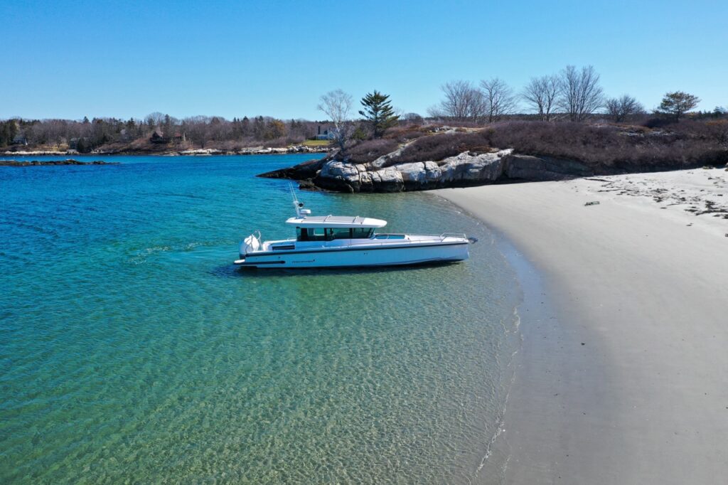 Drone view of an Axopar 37 XC Cross Cabin resting in crystal-clear shallow water alongside a sandy beach and rocky shoreline.