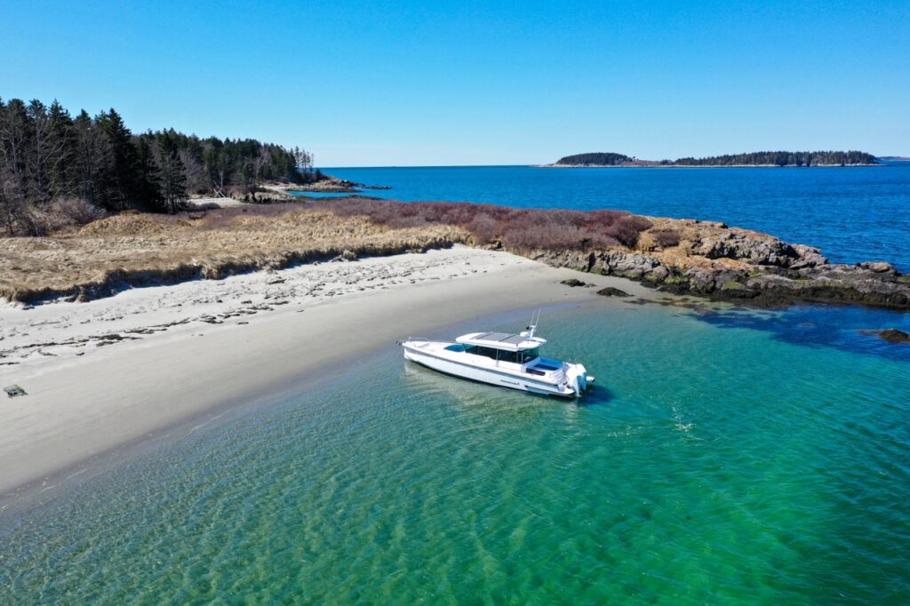 Drone photo of an Axopar 37 XC Cross Cabin in shallow turquoise water just off a secluded sandy beach with rocky shoreline and open ocean beyond.
