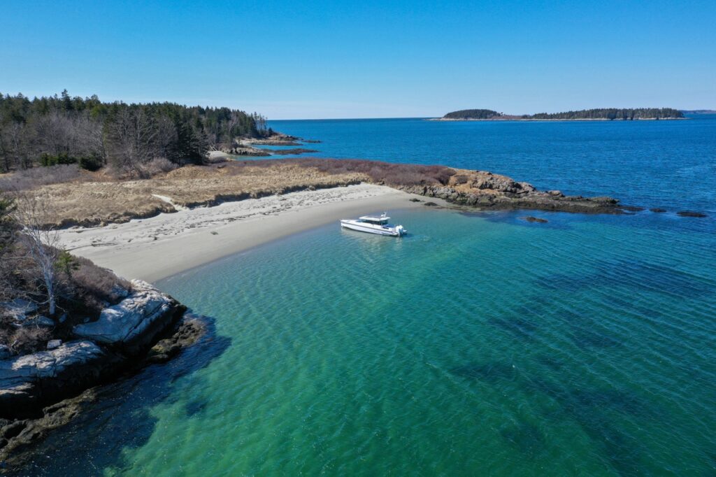 Drone view of an Axopar 37 XC Cross Cabin sitting in clear green-blue water off a sandy cove with rocky shoreline and islands on the horizon.