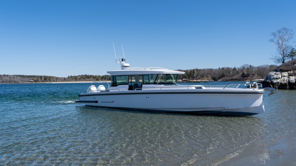 Side view of a white Axopar 37 XC Cross Cabin in clear shallow water beside a sandy beach on a calm, sunny day.