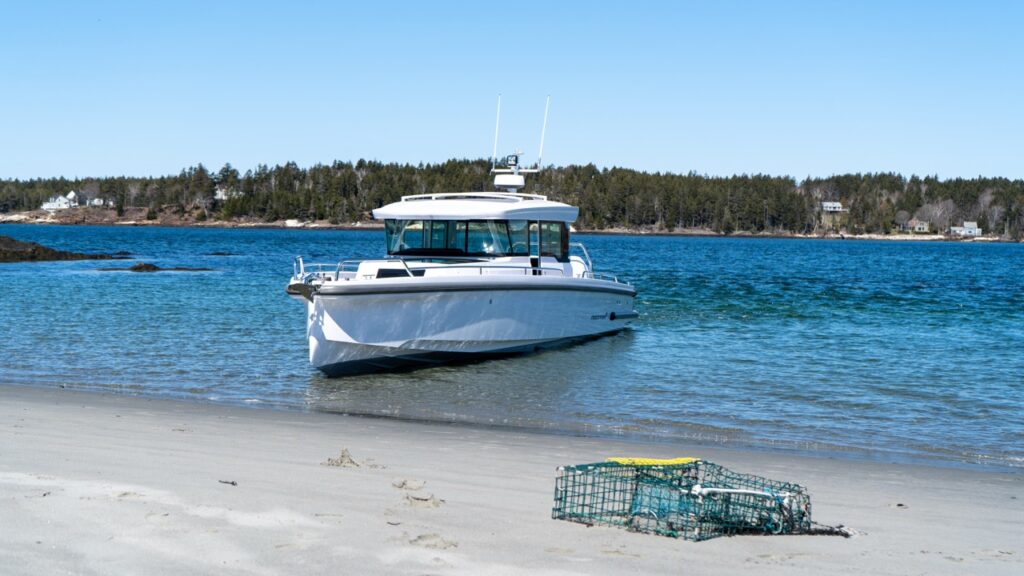 White Axopar 37 XC Cross Cabin beached bow-first near a sandy shoreline with a lobster trap in the foreground and calm blue water behind.