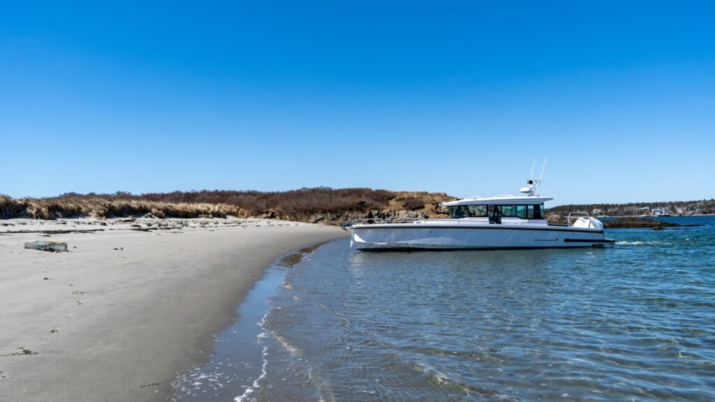 Axopar 37 XC Cross Cabin floating just off a sandy beach in shallow water, viewed from shore with gentle waves and a clear blue sky.
