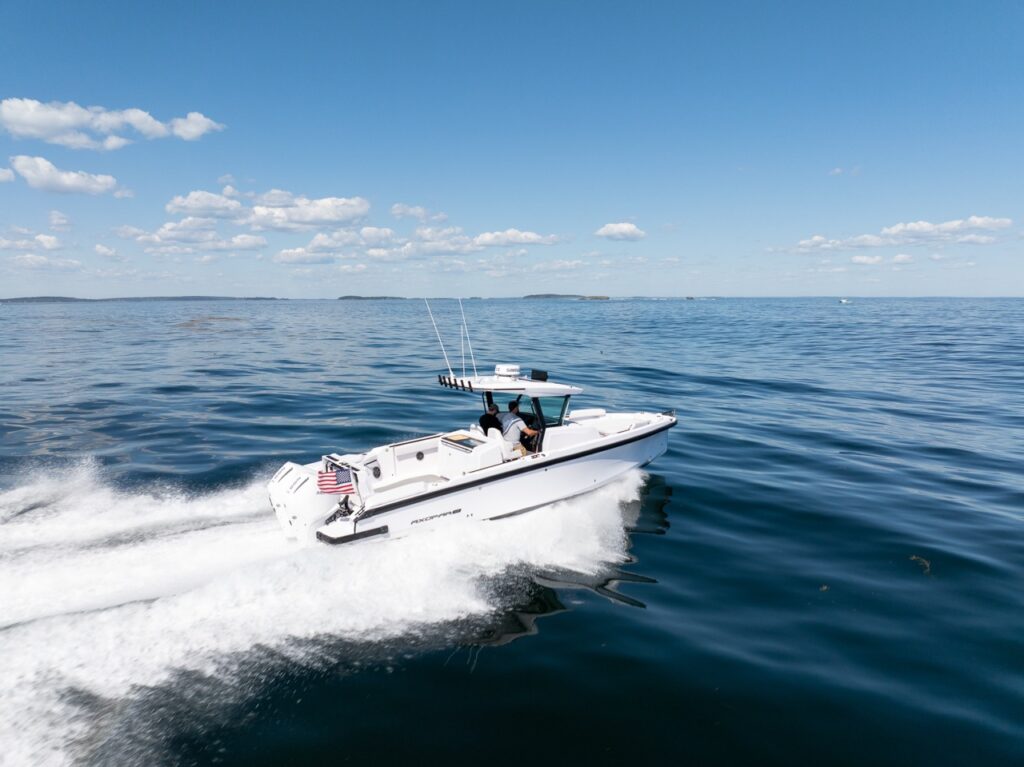 Aerial view of an Axopar 29 CCX center console running on plane across calm open water with a wide white wake, featuring the Cross Top and twin outboards.