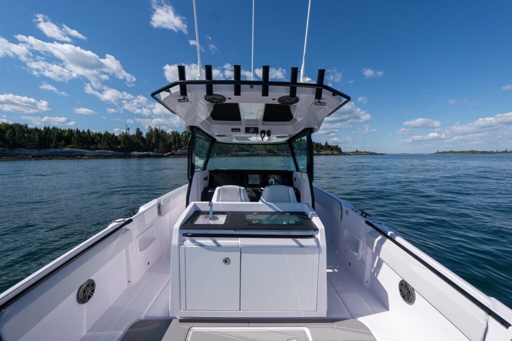Wide cockpit view of the Axopar 29 CCX showing the center-console helm, overhead Cross Top with rod holders, and the aft tackle station featuring a sink and livewell, all set against calm coastal water.