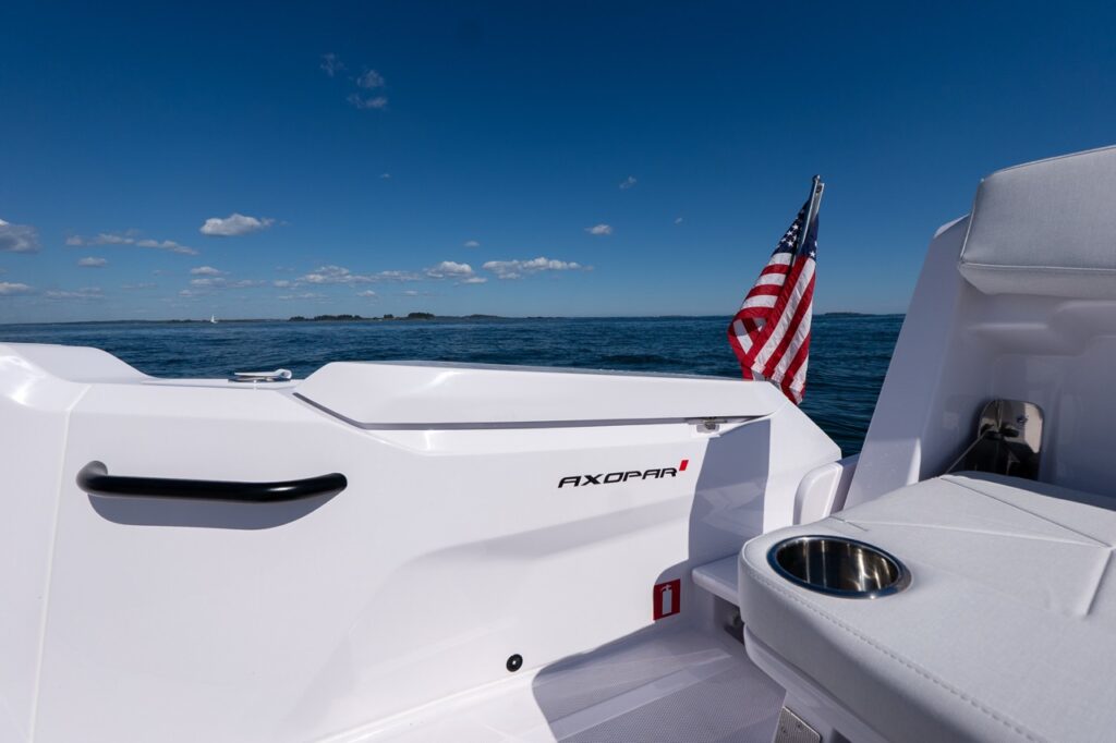 Aft cockpit view of an Axopar 29 CCX showing the Axopar logo, American flag, cupholder, and clean white deck against open water and a clear sky.