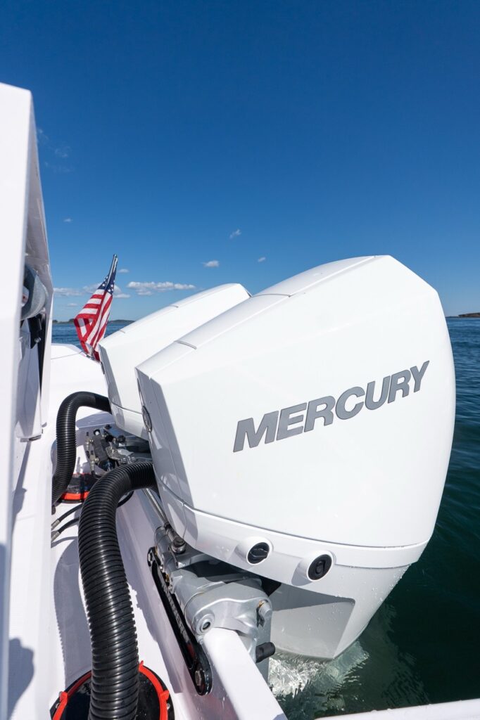 Close-up of two white Mercury Verado outboard engines mounted on the transom of an Axopar 29 CCX, with an American flag and calm water in the background.
