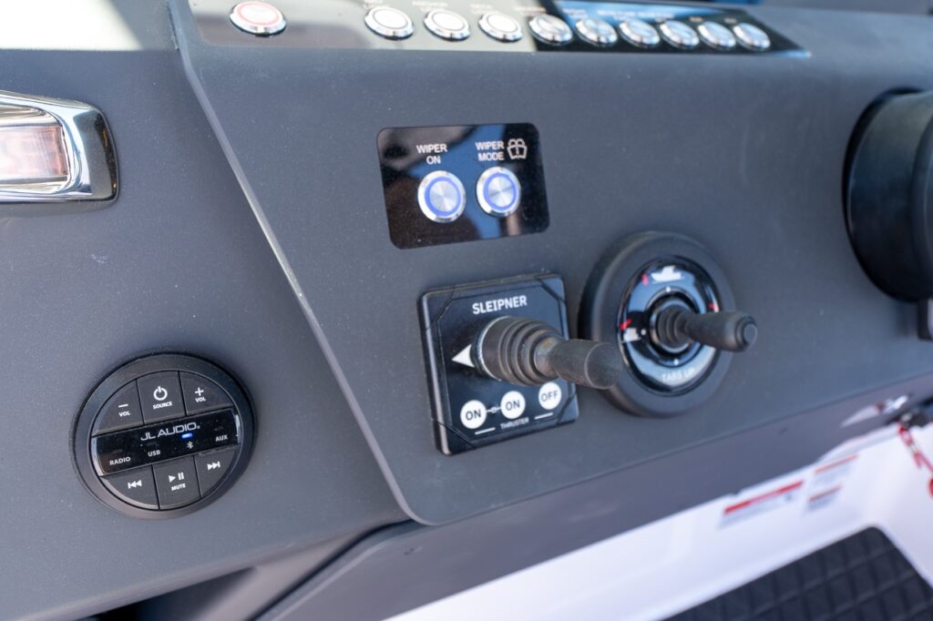 Close-up of the Axopar 29 CCX center console helm showing Sleipner bow thruster joystick, windshield wiper controls, JL Audio remote, and ignition switch panel.