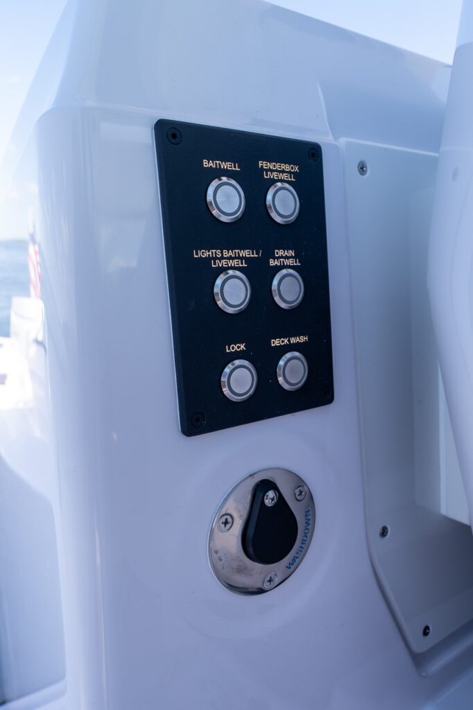 Close-up of an Axopar 29 CCX cockpit switch panel with labeled controls for baitwell, livewell, lights, drain, lock, and deck wash, above a washdown seacock valve.