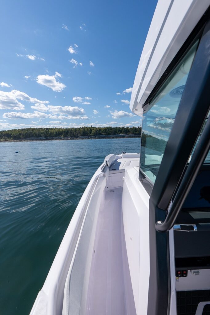 Starboard-side walkaround deck on an Axopar 29 CCX center console, looking forward along the gunwale with calm coastal water and a tree-lined shoreline in the background.