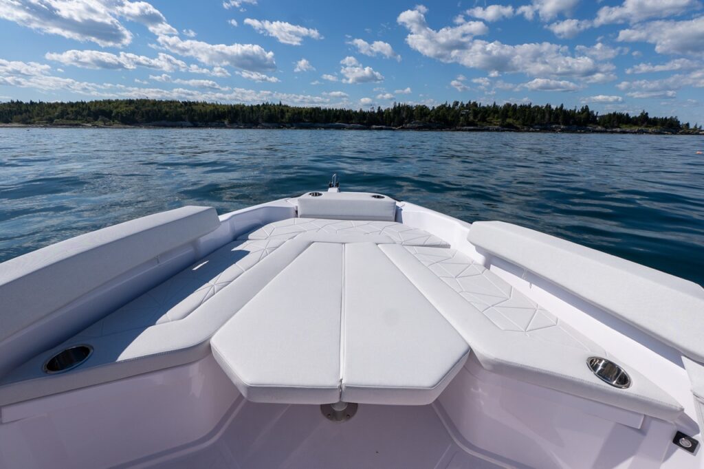 Axopar 29 CCX center console bow lounge with a large white sun pad, wraparound seating, and built-in cupholders, looking out over calm water and a wooded shoreline.