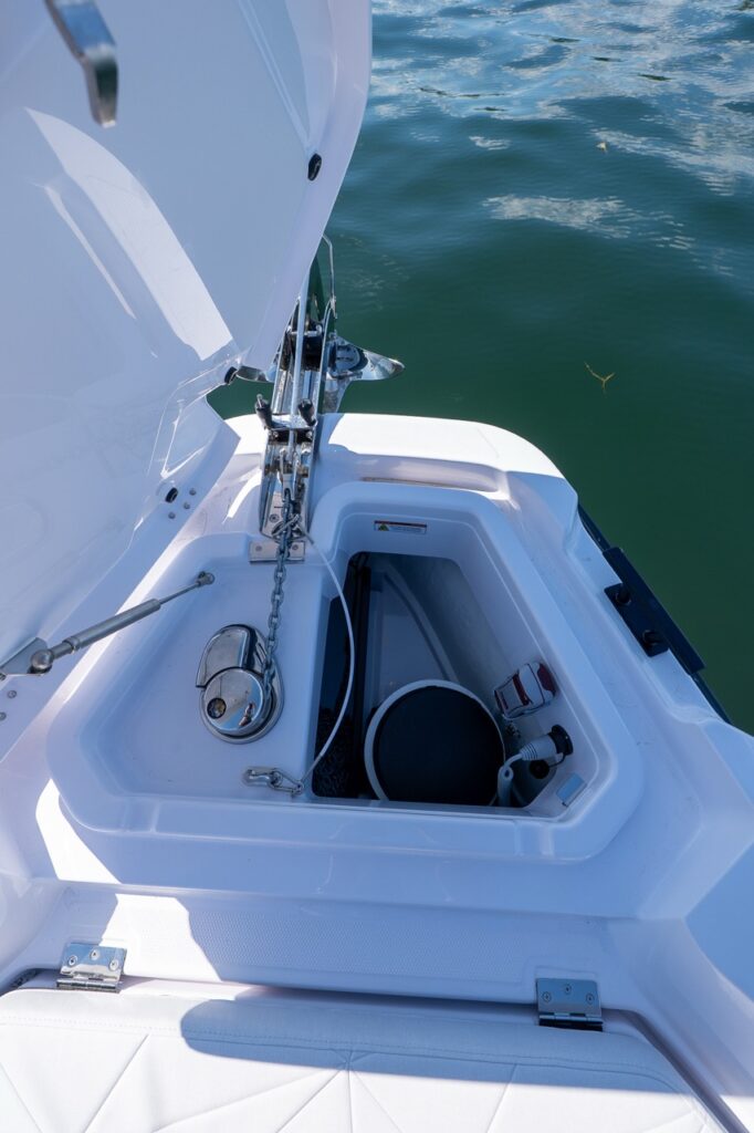 Open bow anchor locker on an Axopar 29 CCX center console showing the anchor rode/chain, windlass hardware, and deep storage compartment with calm water alongside.