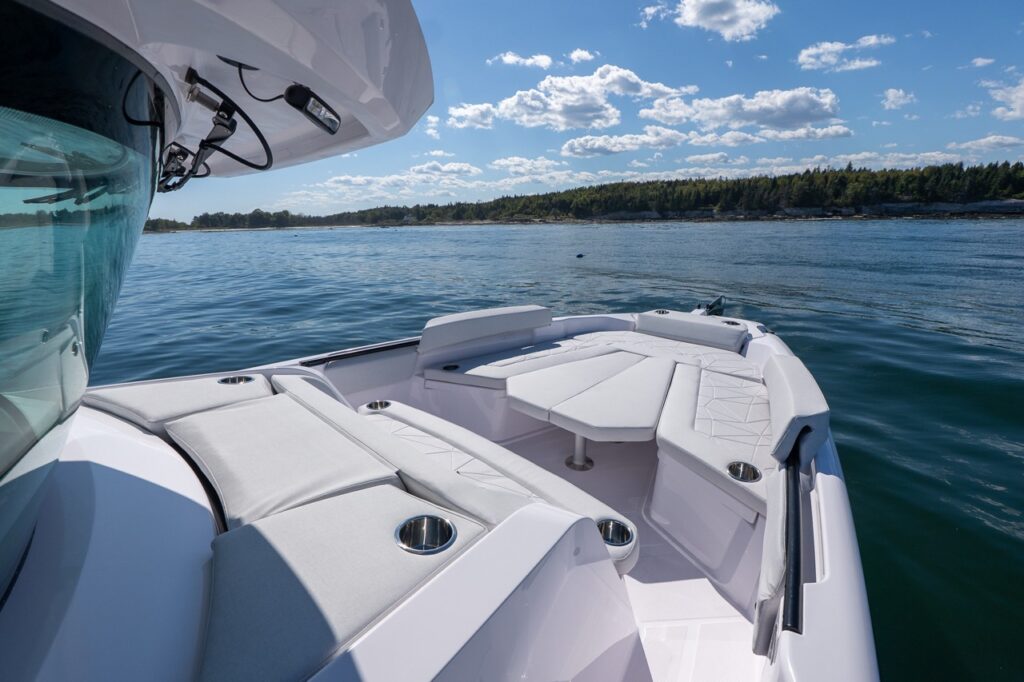 Bow seating and sun pad on an Axopar 29 CCX center console, showing wraparound forward lounge, removable table, and cupholders with calm water and a tree-lined coastline.