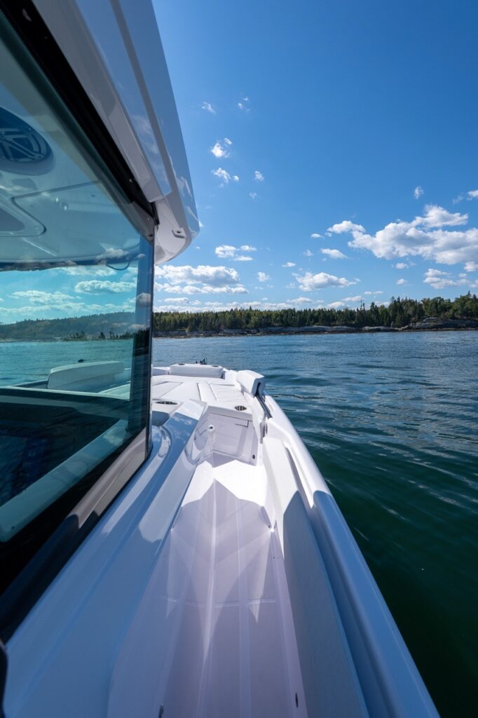 Starboard-side walkaround deck on an Axopar 29 CCX center console, leading forward to the bow seating area with calm water and a wooded shoreline in the background.