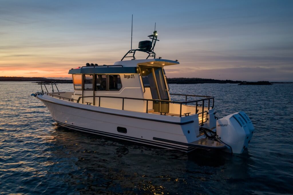 Aft three-quarter view of a Targa 27.2 Coupe at sunset, highlighting the enclosed pilothouse, extended roof overhang, secure aft deck with railings, and twin Mercury outboards.