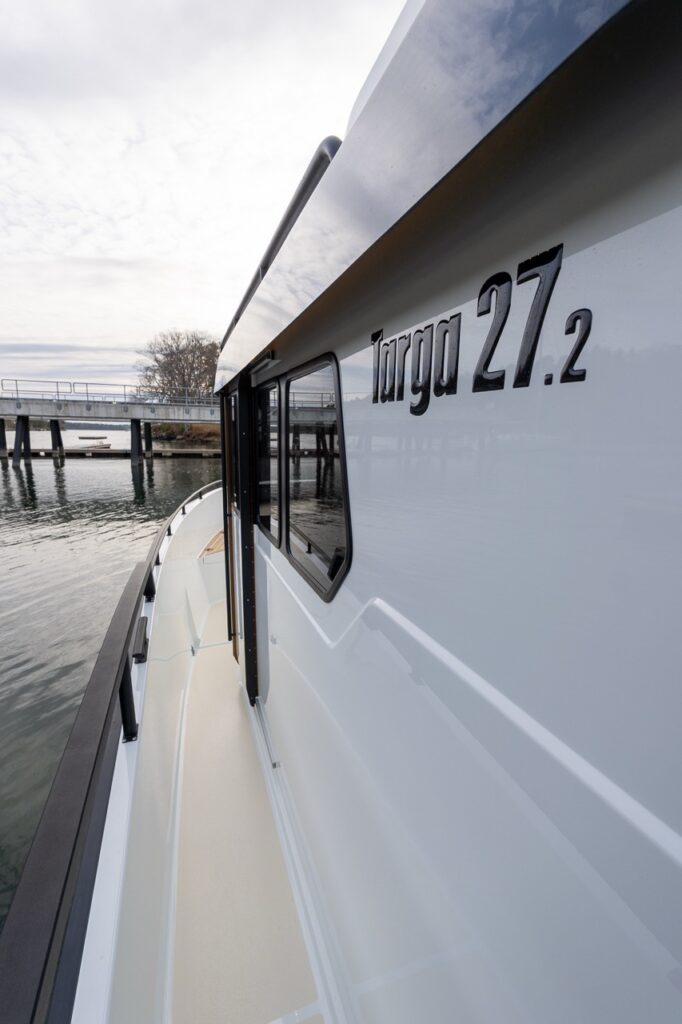 Close-up of the Targa 27.2 Coupe side deck at the dock, showing the wide walkaround passage, non-skid deck surface, tall safety rail, pilothouse side windows, and raised Targa 27.2 hull lettering.