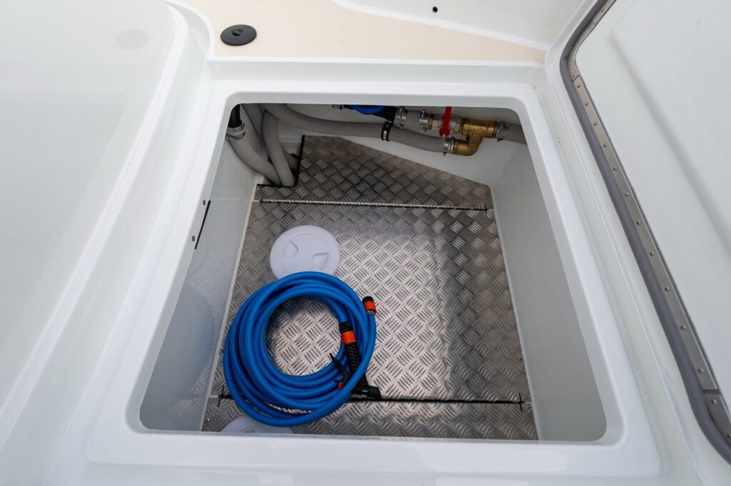 Open below-deck storage compartment on a Targa 27.2 Coupe, showing a deep molded space with aluminum tread plate flooring, freshwater hose, plumbing connections, and clean structural finishes.