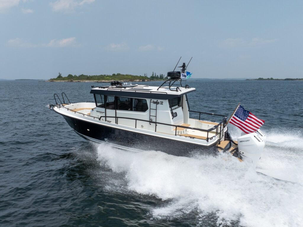 A Targa 32 pilothouse boat running at speed on open water, viewed from aft-quarter, with twin Mercury outboards creating a strong wake and an American flag flying at the stern.