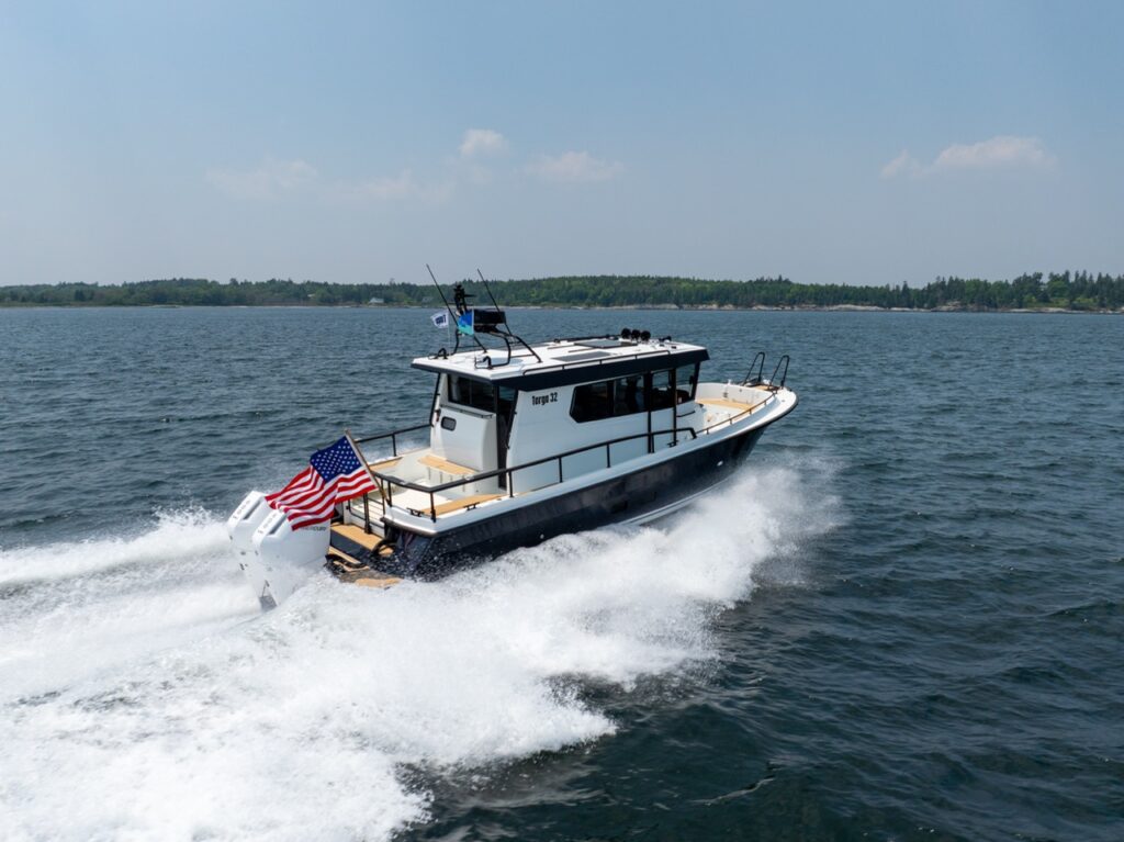 A Targa 32 pilothouse boat running at speed on open water, viewed from above and aft, with twin Mercury outboards creating a wide wake and an American flag flying at the stern.