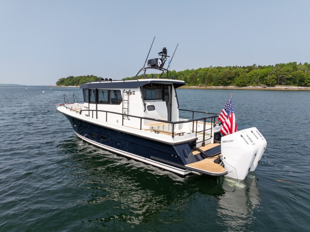 Three-quarter stern view of a Targa 32 pilothouse boat at rest on calm coastal water, showing the aft deck, American flag, teak swim platform, and Mercury outboard power.